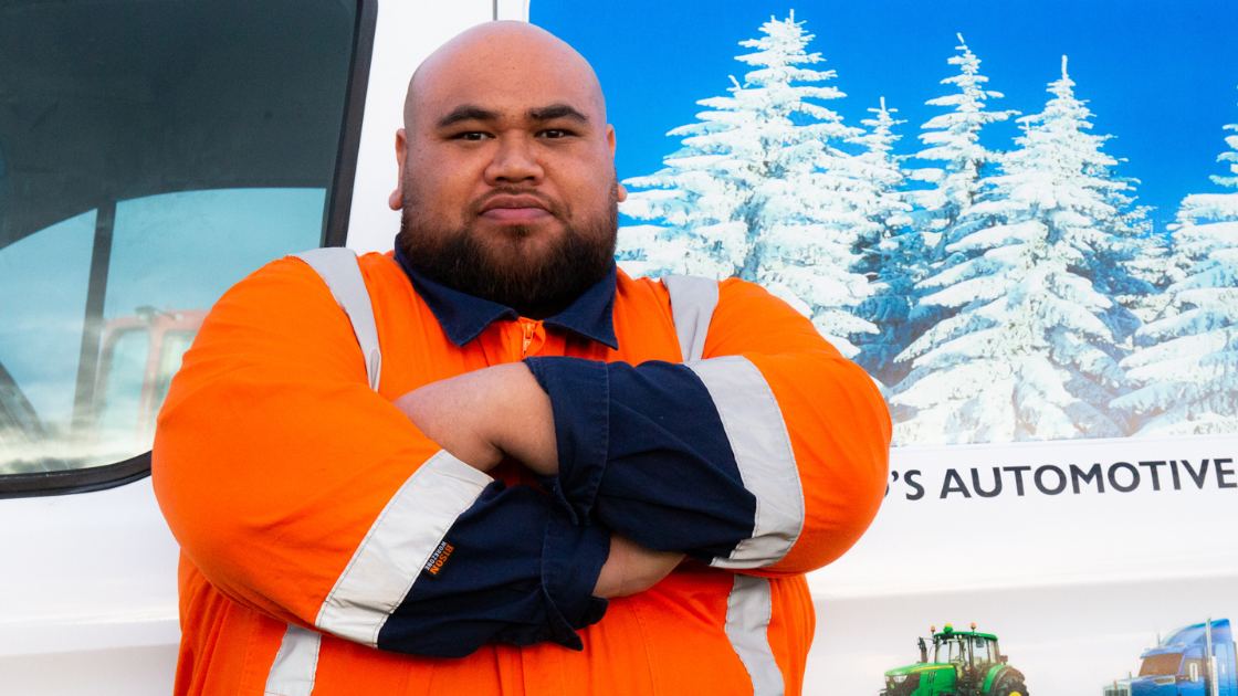 A worker in hi vis overalls standing with arms folded beside a truck, looking straight at the camera